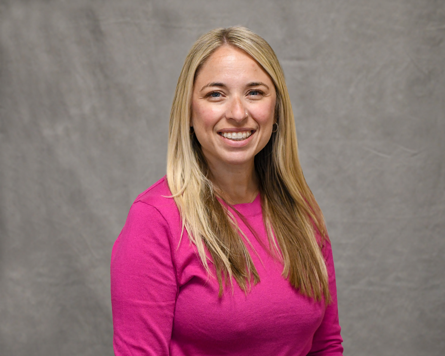 Blonde woman in a pink sweater smiling, posing in front of a grey backdrop.
