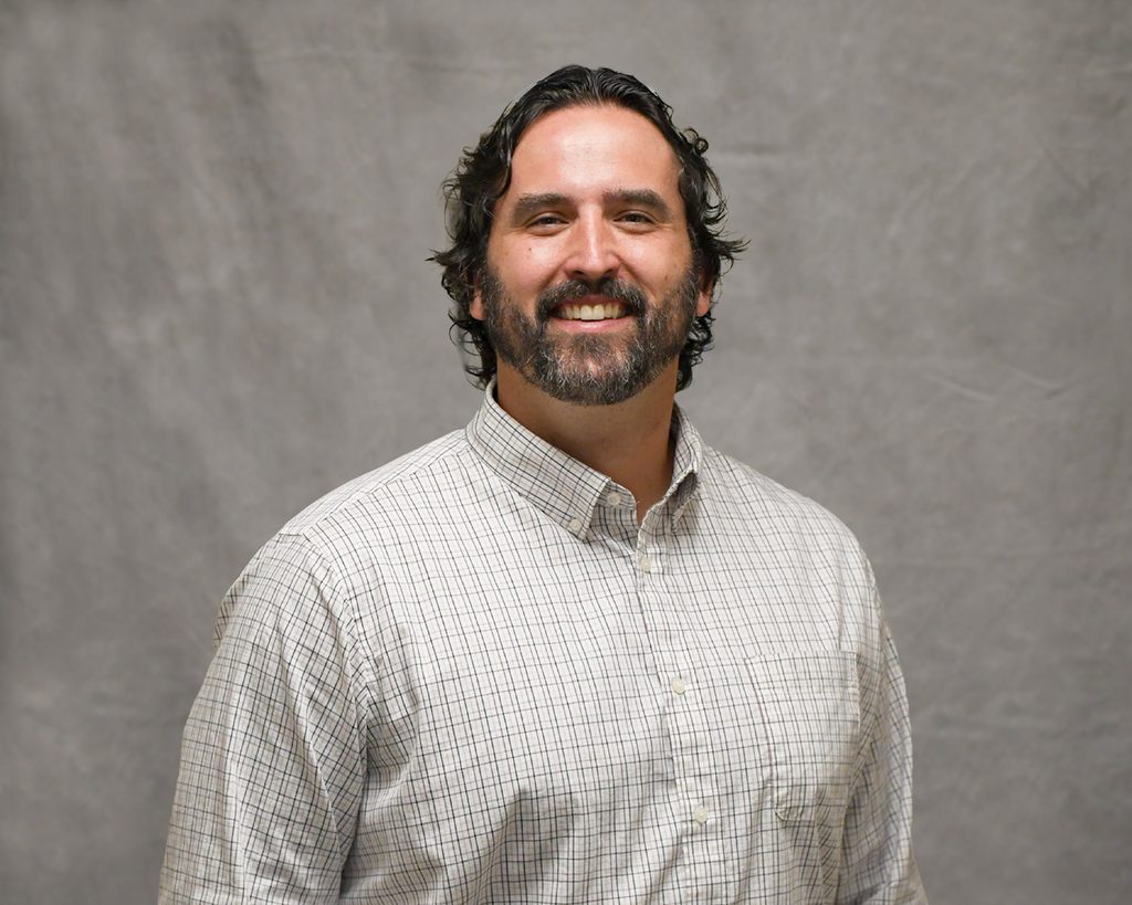 Male health care provider with dark hair and beard smiling wearing a long sleeve button down checkered shirt.