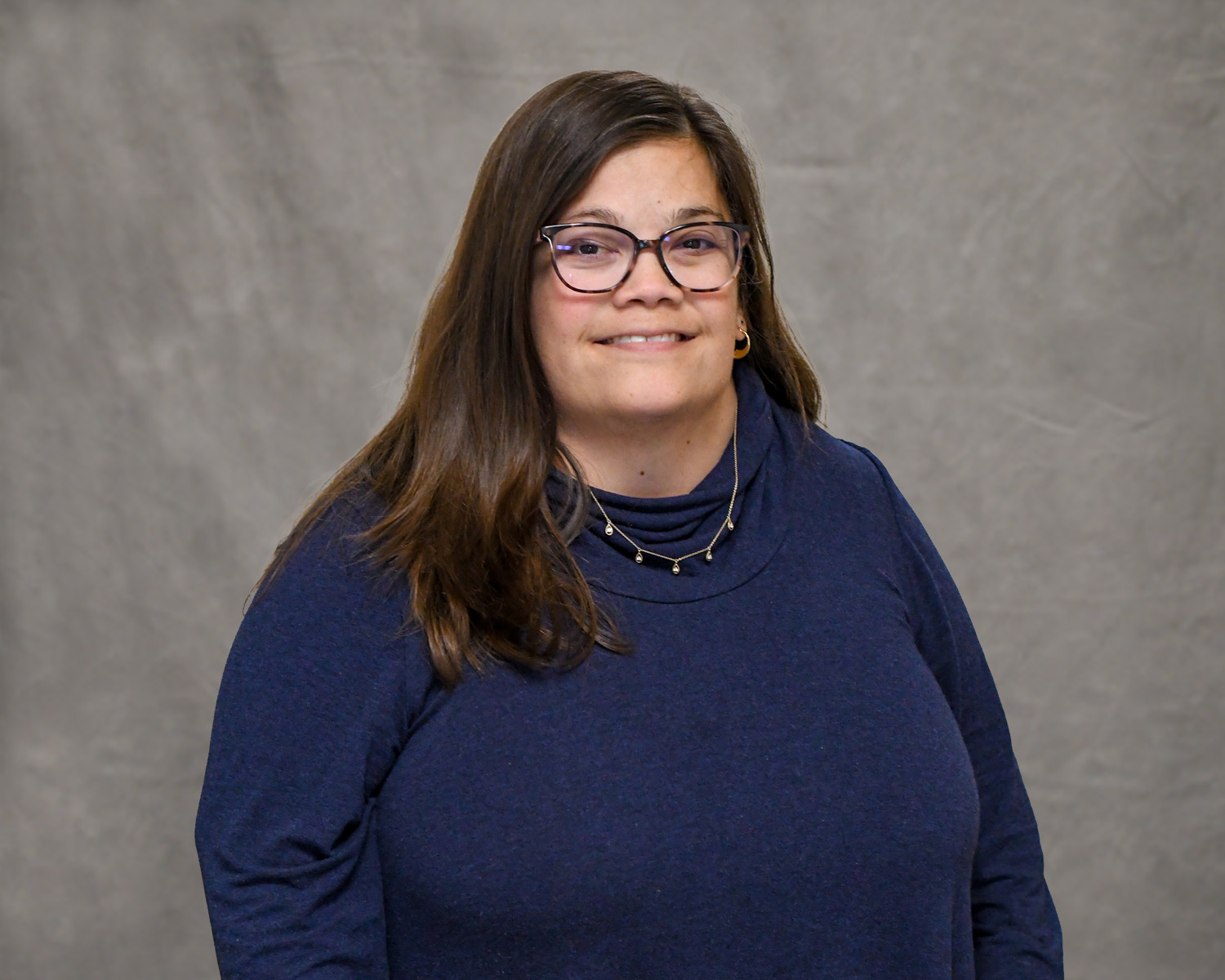 A Caucasian woman in a navy sweater with long black hair poses for the camera in front of a grey backdrop.